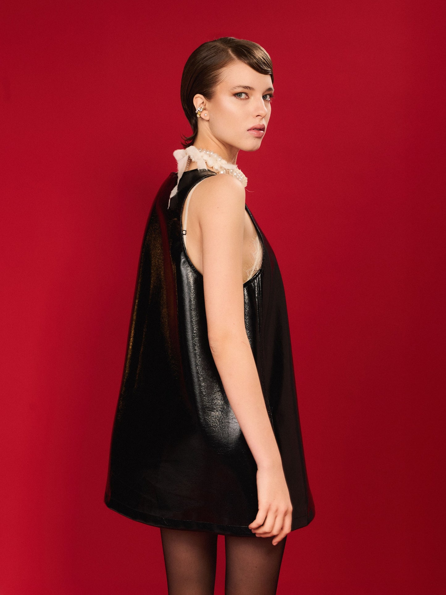 Black high-gloss A-line mini dress with deep scoop neckline and sleeveless cut, styled with a pearl necklace - TeAdore.